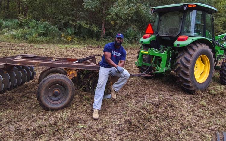 Christopher Joe, AL, wearing a blue t-shirt that says “Make Farming Great Again” and light blue jeans leaning on tractor equipment.
